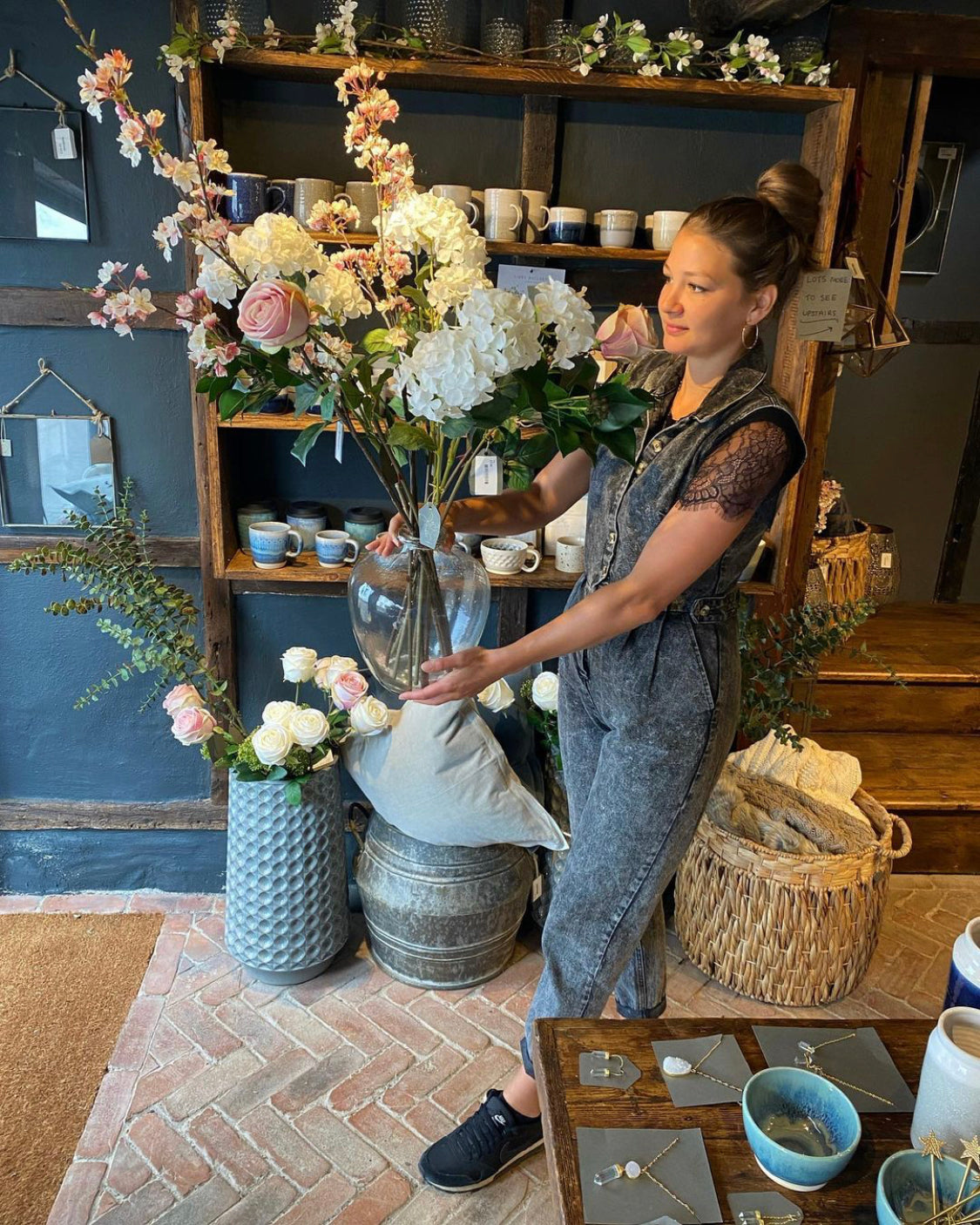 Rose and Co store interior in Godalming. Woman holding large bouquet of fake, artificial flowers in glass vase surrounded by mugs, photo frames, jewellery, baskets and other homeware items