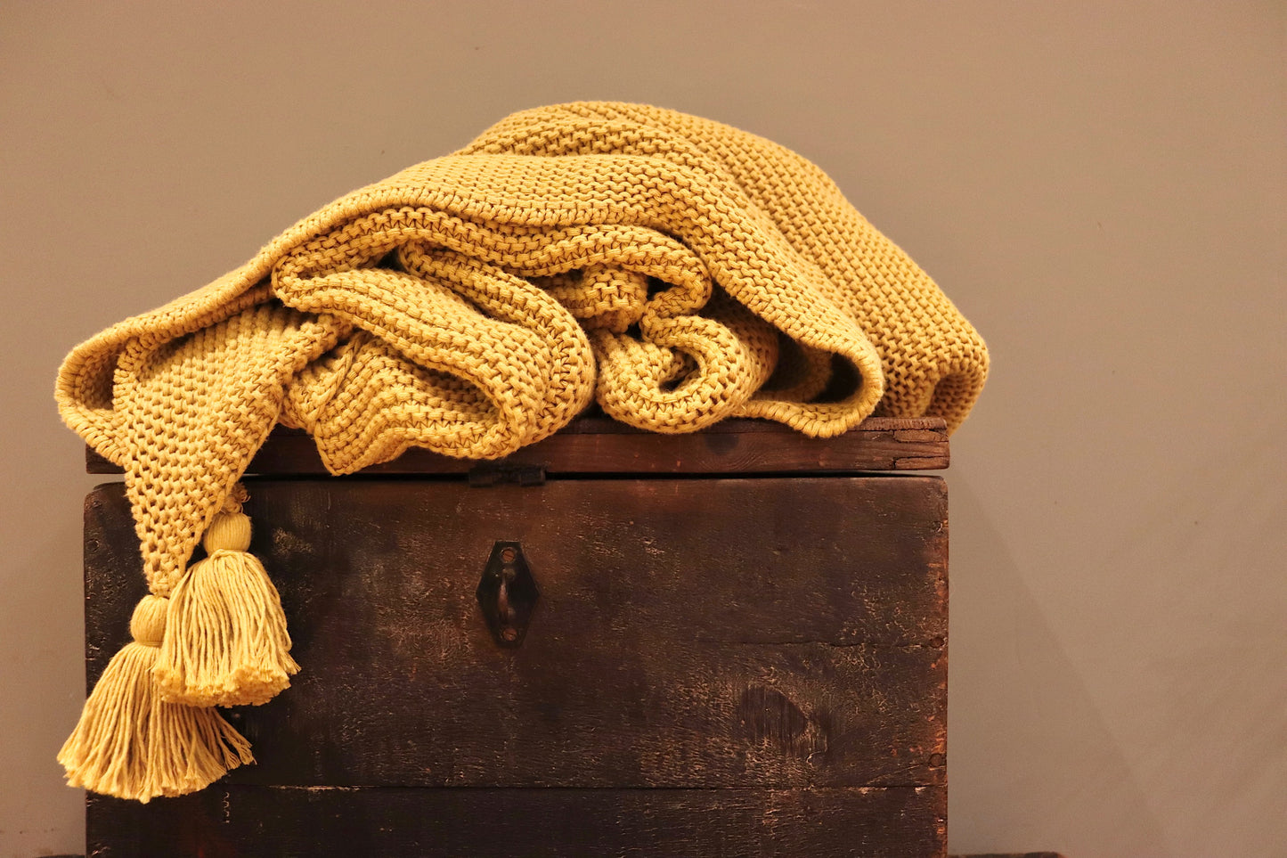 Mustard yellow chunky knit throw with 4 large tassels at each corner. The knitted throw is sitting on top of a small dark wooden chest.