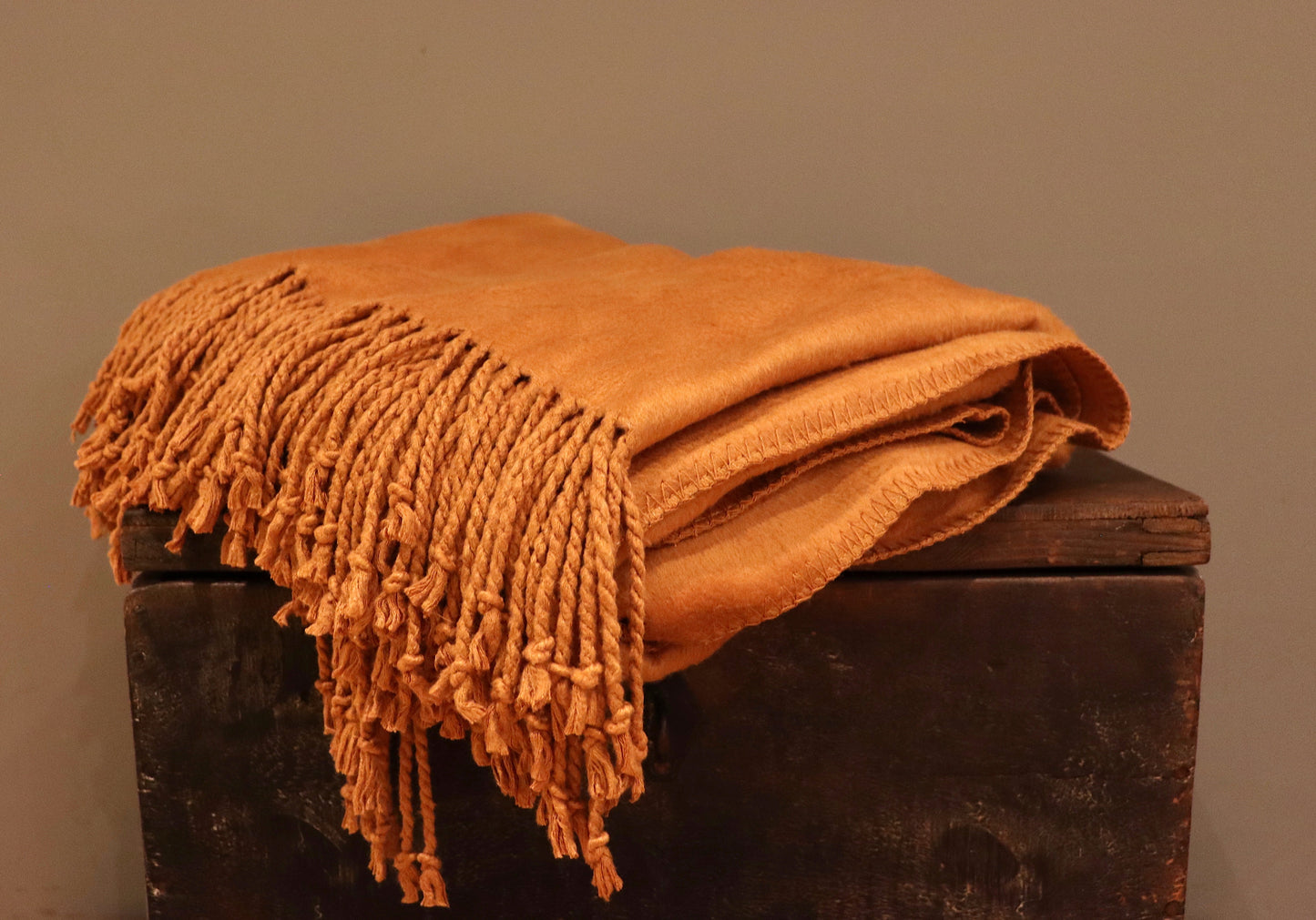 Burnt orange bamboo throw, with a tassel fringe, folded up on a dark wood trunk.