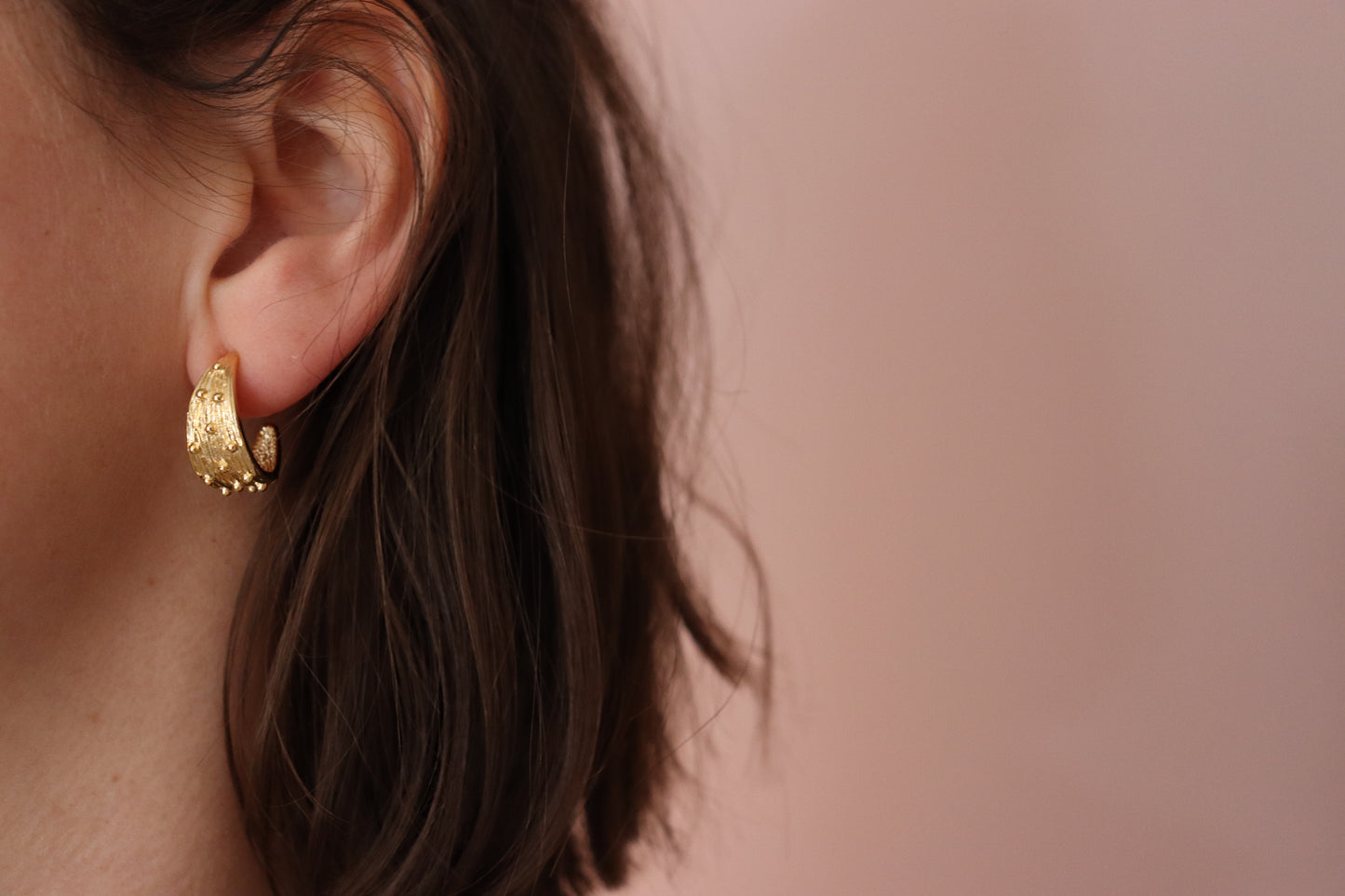 Gold Plated Half Hoop Earring, with little spikes covering its half moon shape. Worn on a left ear, with brown hair and a pink background.