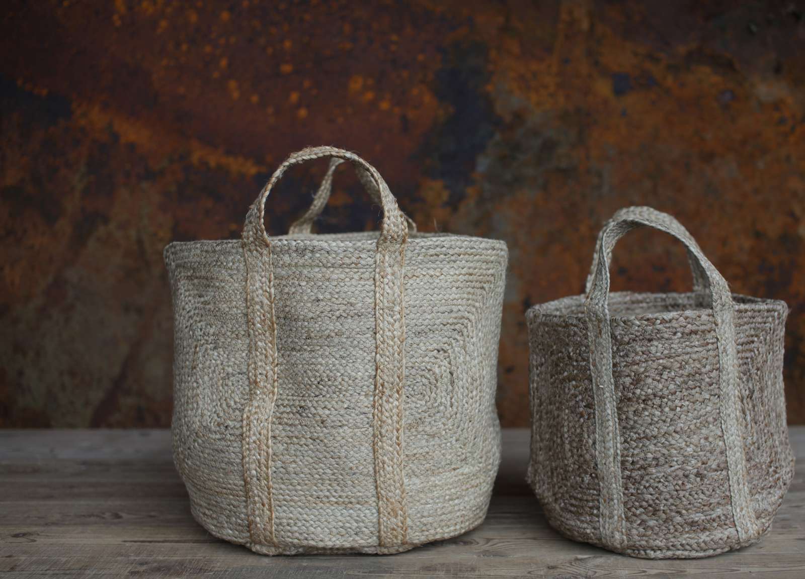 Two natural, braided hemp storage bags. One Large, one small, with handles, standing on a wooden surface with a rusty background.