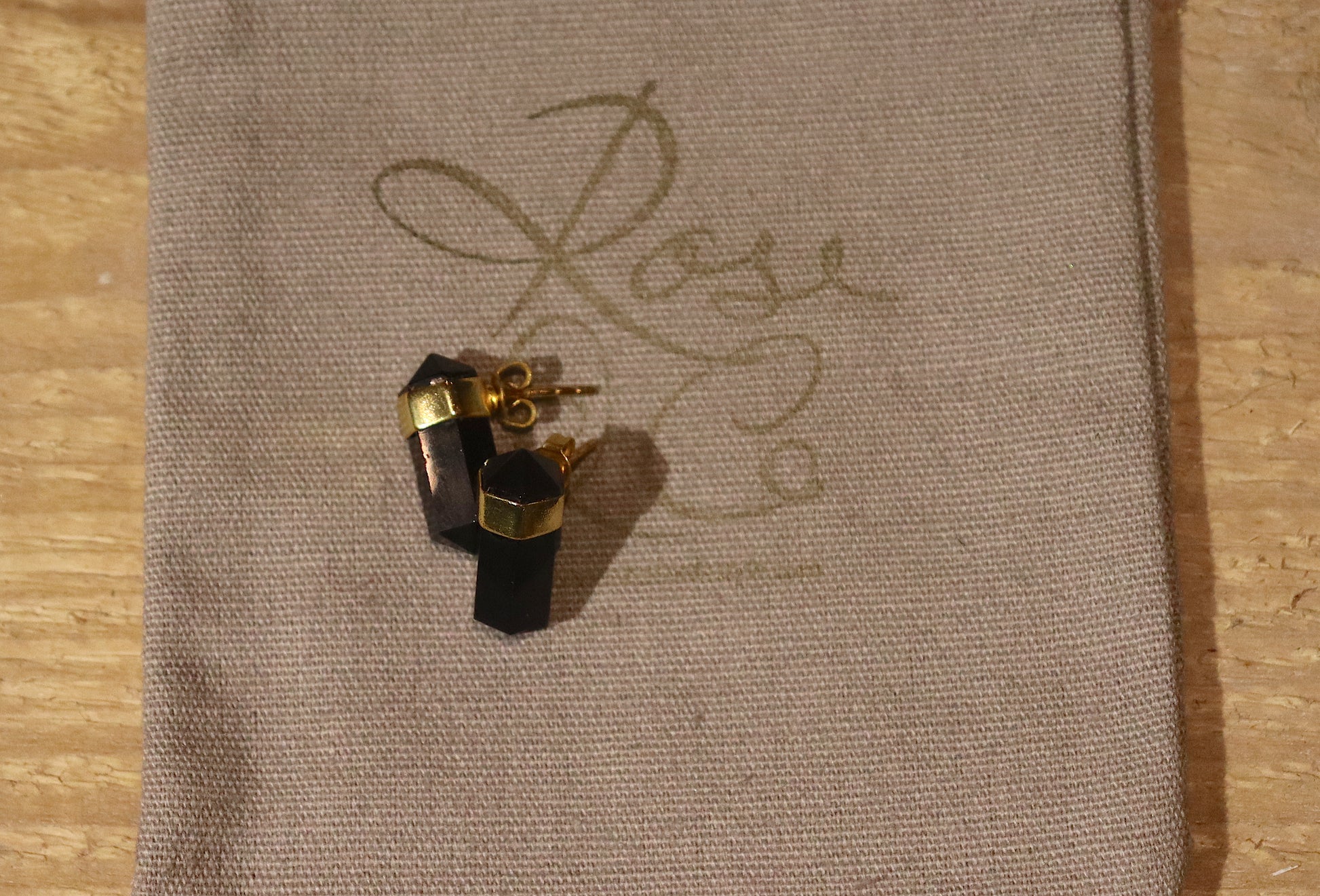 A pair of black crystal stud earrings in a gold plated setting on a grey cotton drawstring bag with a gold rose and co label stamped onto it.