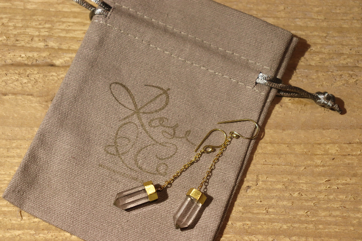 Gold plated clear quartz crystal points on a dangly chain, on a grey cotton drawstring bag with a gold Rose & Co logo stamped on it.