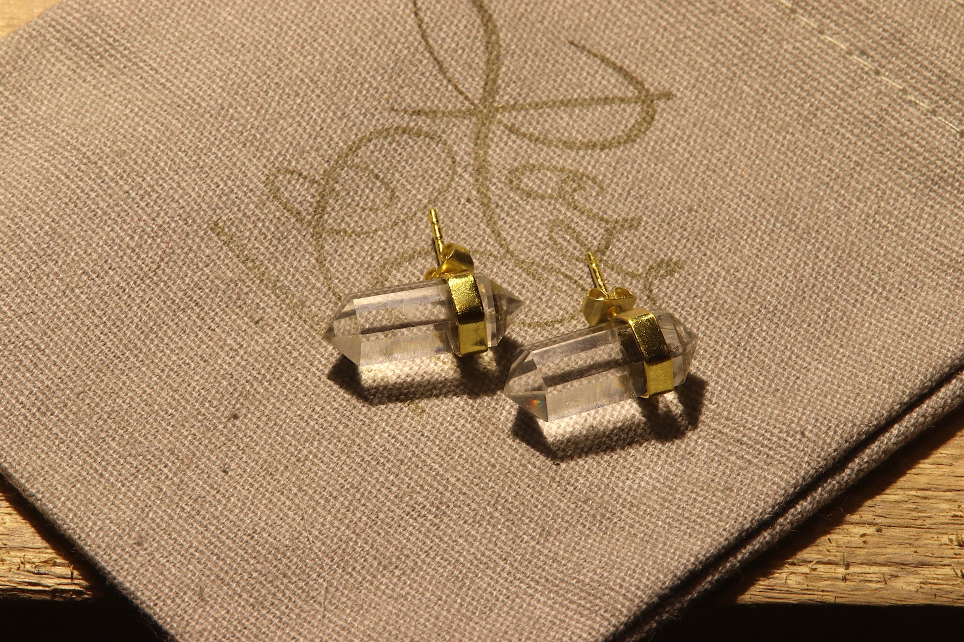 Clear crystal earrings in gold casing on a grey cotton drawstring bag with a gold rose and co logo stamped on it.