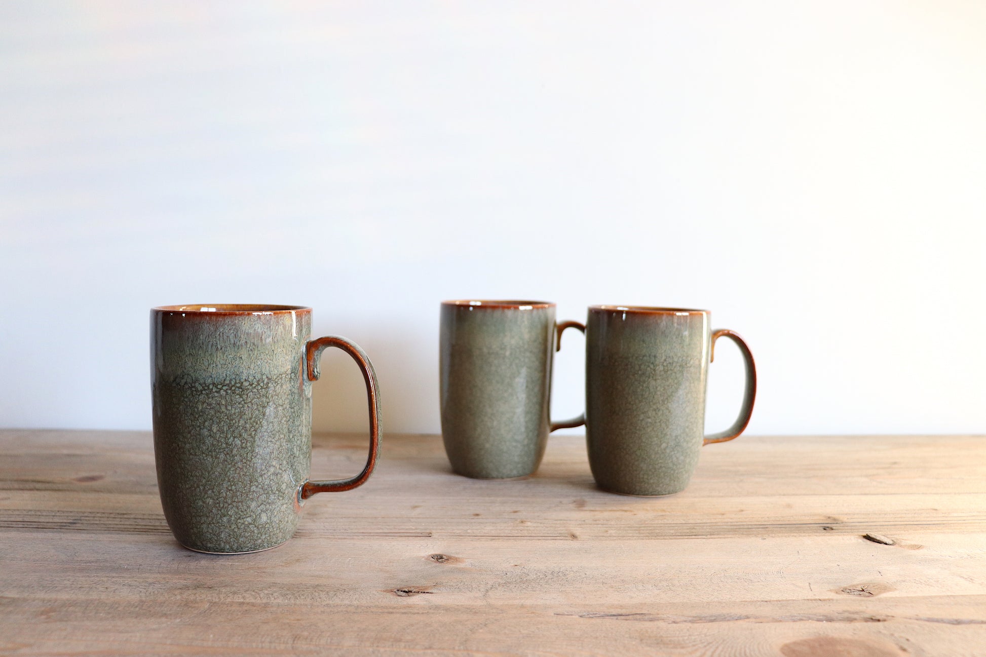 Tall green mug with a brown rim and edge of handles. The green glaze is slightly iridescent , giving a beautiful overall effect. The colour is both on the outside and inside of the cup. Standing on a wooden surface with white background.