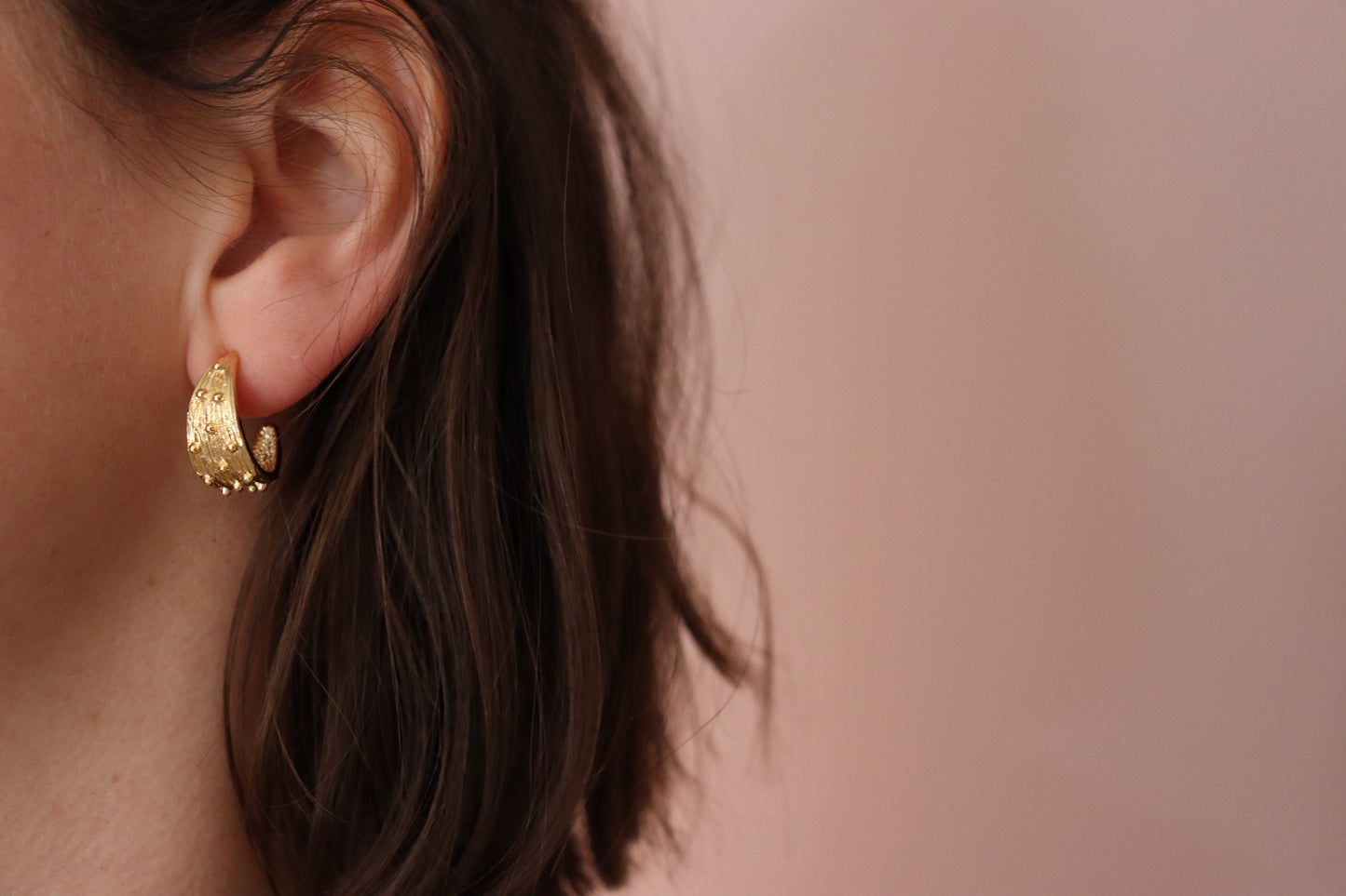 Gold Plated Half Hoop Earring, with little spikes covering its half moon shape. Worn on  a left ear, with brown hair and a pink background. 