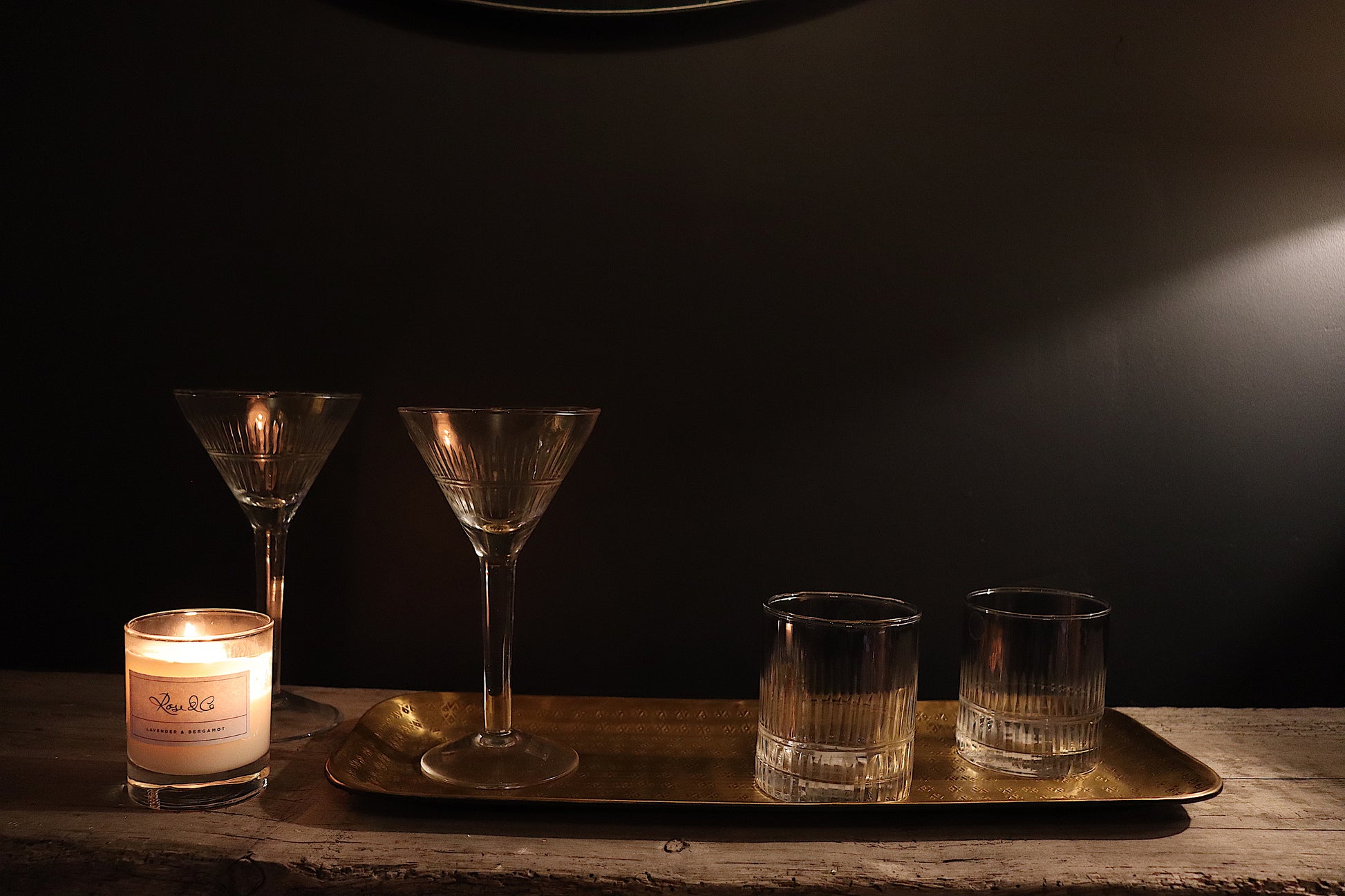 A brass tray with hand made etchings, with two cut-glass cocktail glasses, two cut-glass tumblers with a smokey hue, and a soy candle made with essential oils in a clear glass jar.